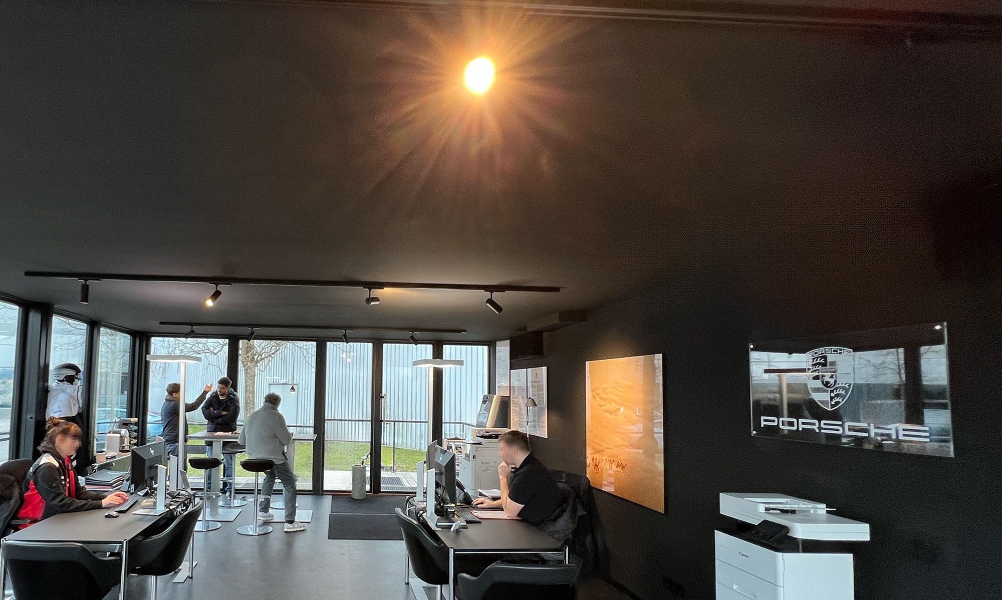 Innenraum eines KOYA Moduls im Porsche Zentrum Aargau: Schreibtische und Hochtische vor einer Glasfront, schwarze Wände, Schienenstrahler