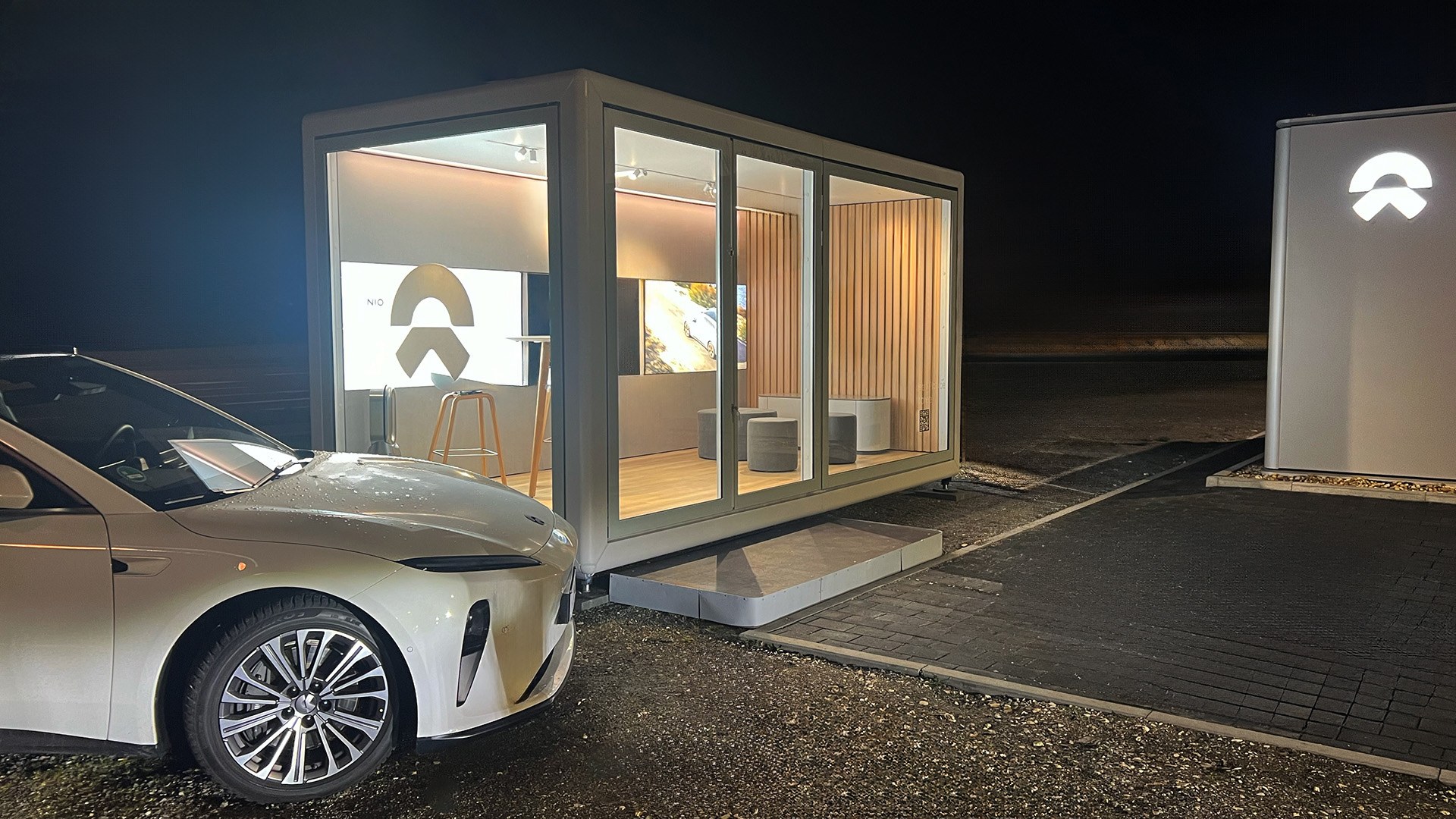 Weißes KOYA Modul mit Glasfront und beleuchtetem Innenraum bei Nacht, davor ein Fahrzeug, rechts eine Stele mit NIO-Zeichen