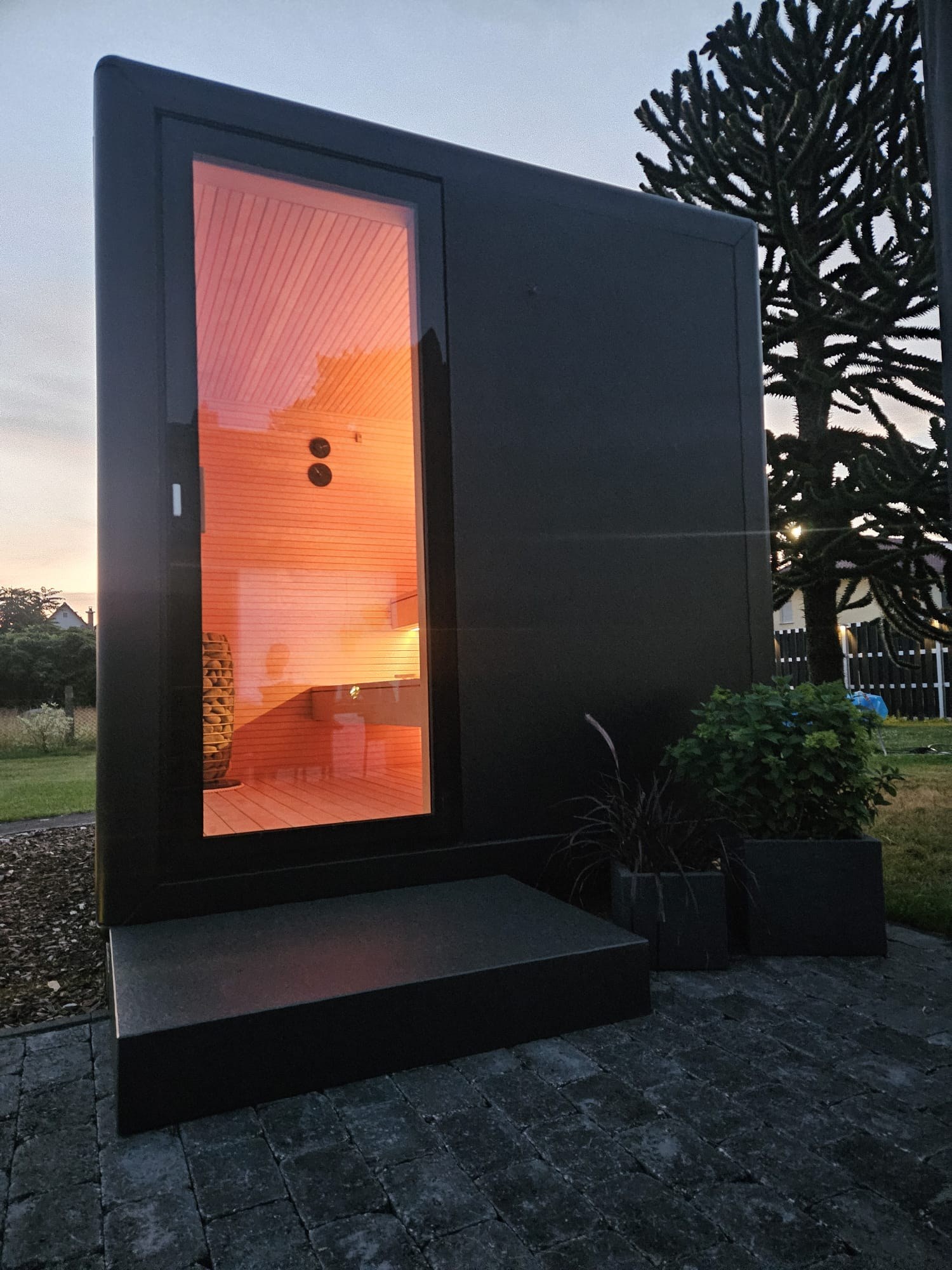Aufnahme: Seitenansicht einer gebauten KOYA Sauna Cube in der Dämmerung, hinter der hohen Glastür warmes Licht