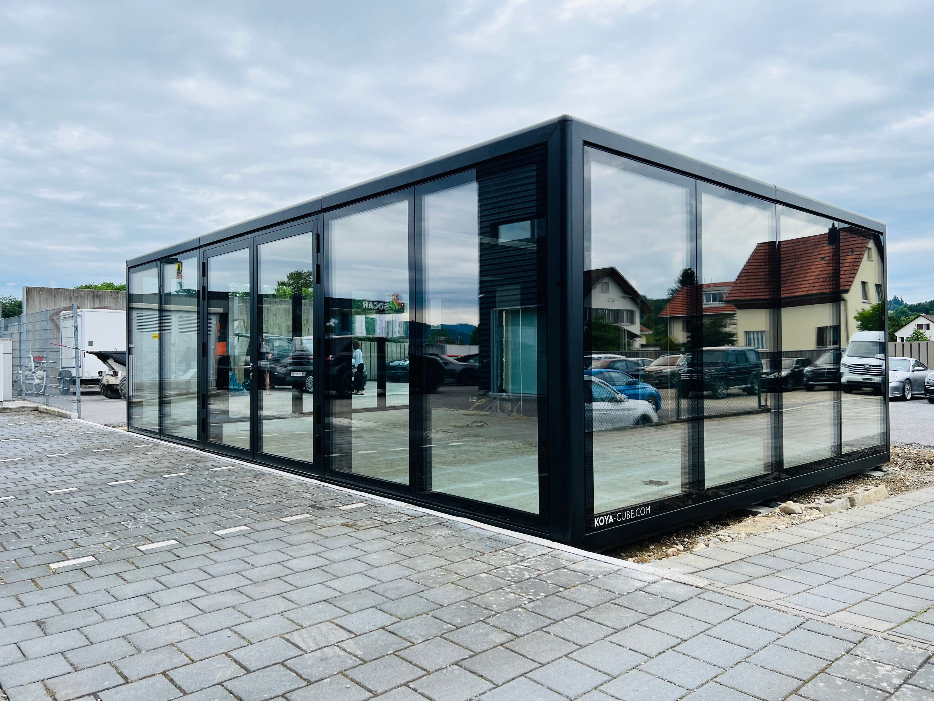 Schwarz gerahmter Glaskubus mit Glastüren auf gepflastertem Platz, Schriftzug KOYA-CUBE.COM an der Glasfront.
