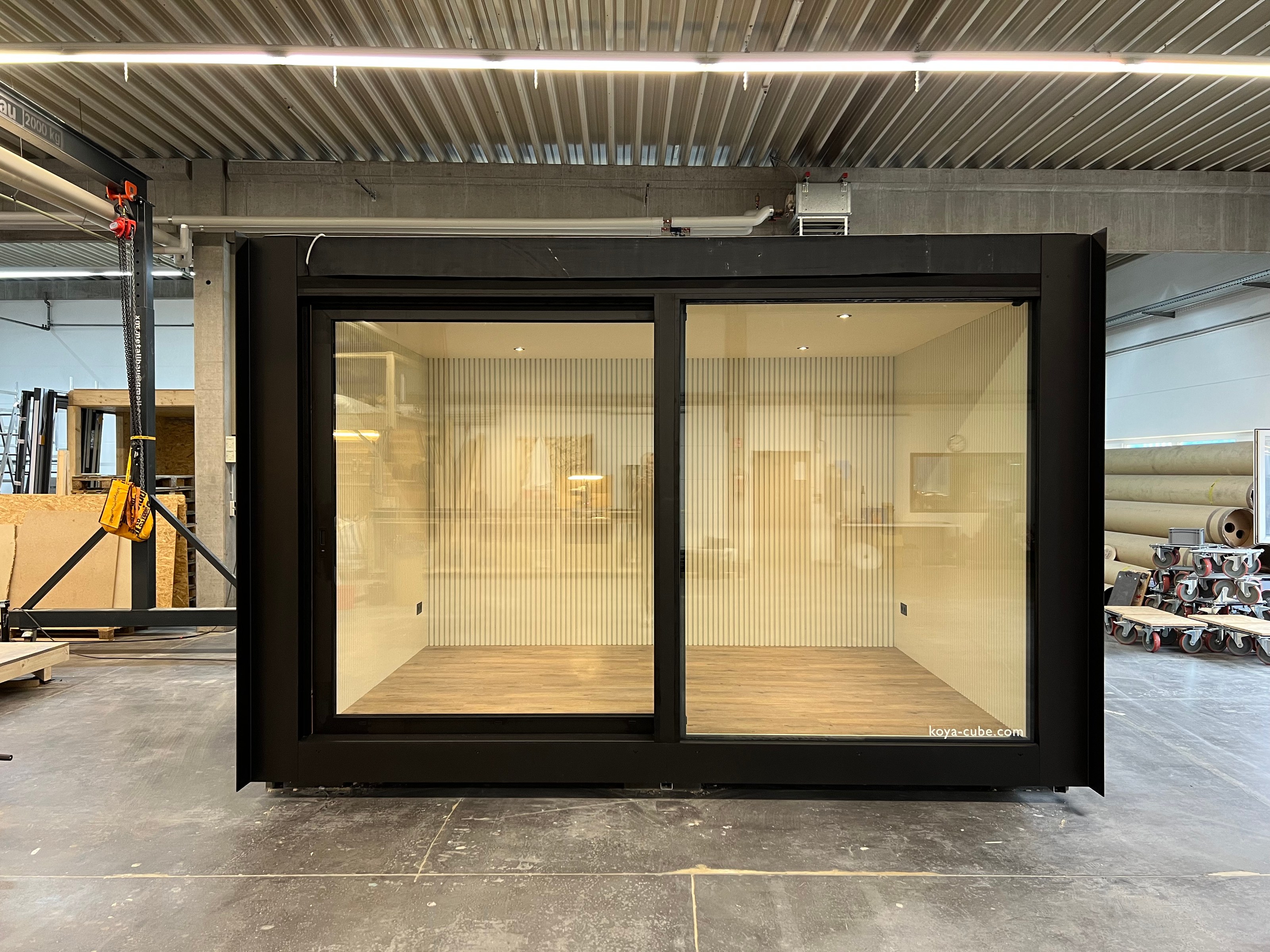 Schwarz gerahmter Glaskubus mit zwei Schiebefronten in einer Halle, innen helle Lamellenwand, Schriftzug koya-cube.com.