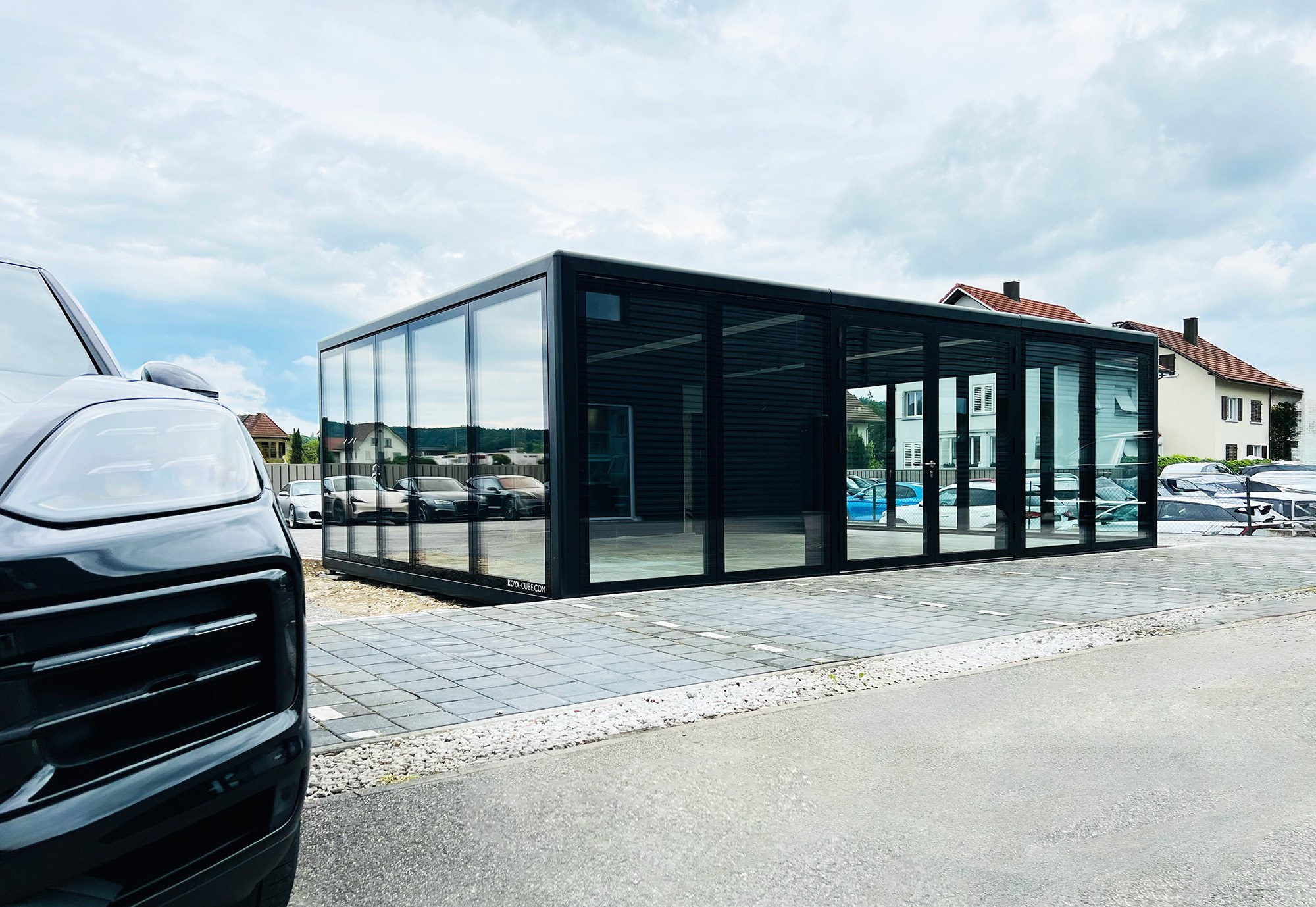 Schwarzer Glaskubus auf gepflasterter Fläche, links die Front eines dunklen Autos, am Sockel der Schriftzug KOYA-CUBE.COM.