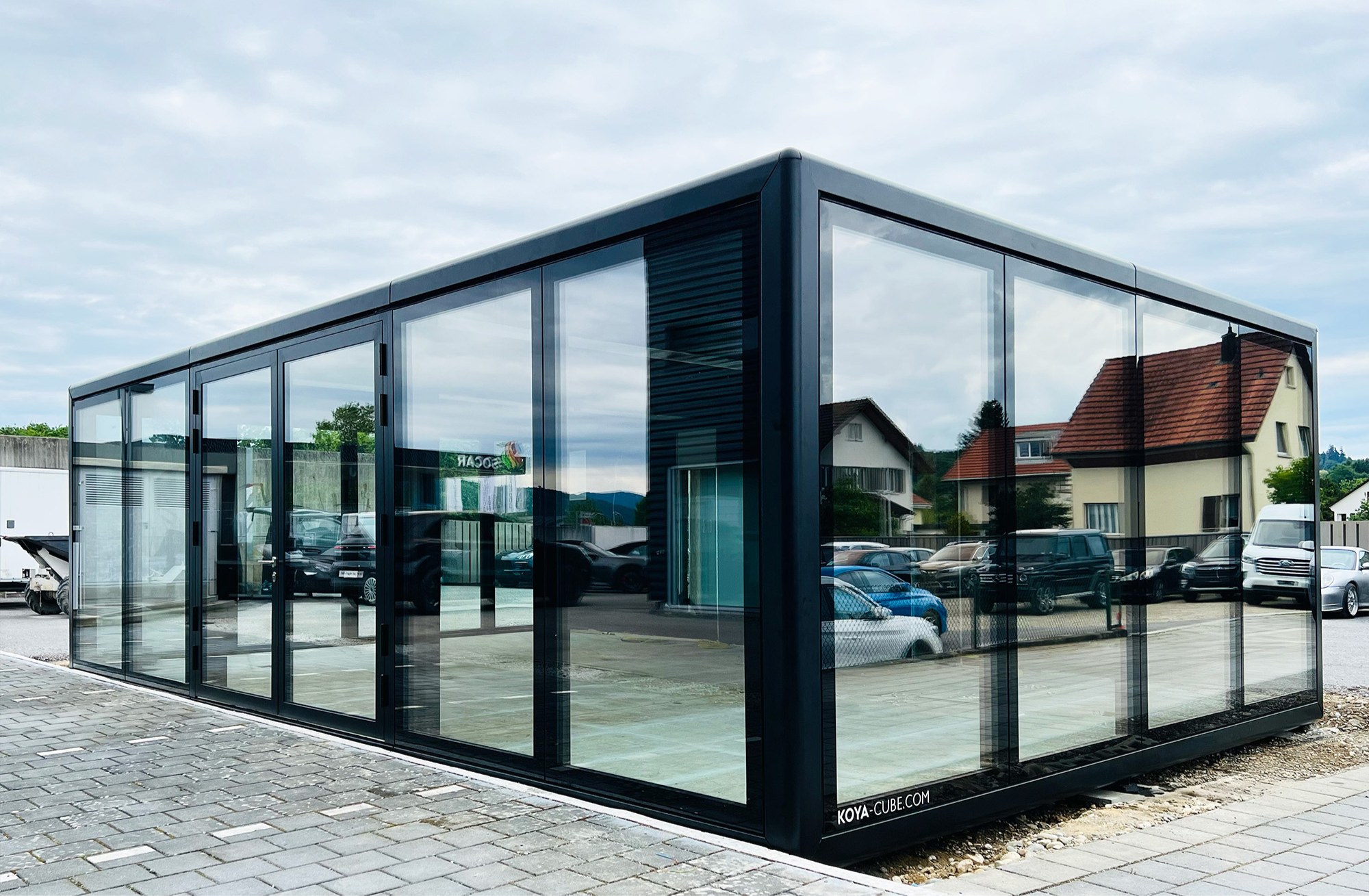 Schwarz gerahmter Glaskubus mit großen Glasfronten und Glastüren auf gepflasterter Fläche, am Sockel KOYA-CUBE.COM.