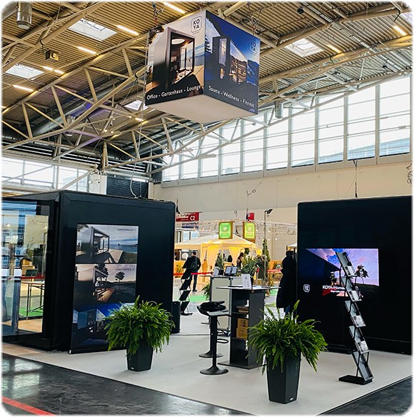 Messestand mit schwarzen Wänden, links ein Glaskubus, von der Decke hängt ein Würfel mit Fotos und KOYA-Logo.