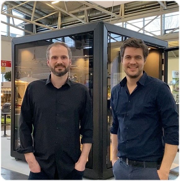 Zwei lächelnde Männer in dunklen Hemden stehen vor einem schwarzen Glaskubus auf einer Messe.