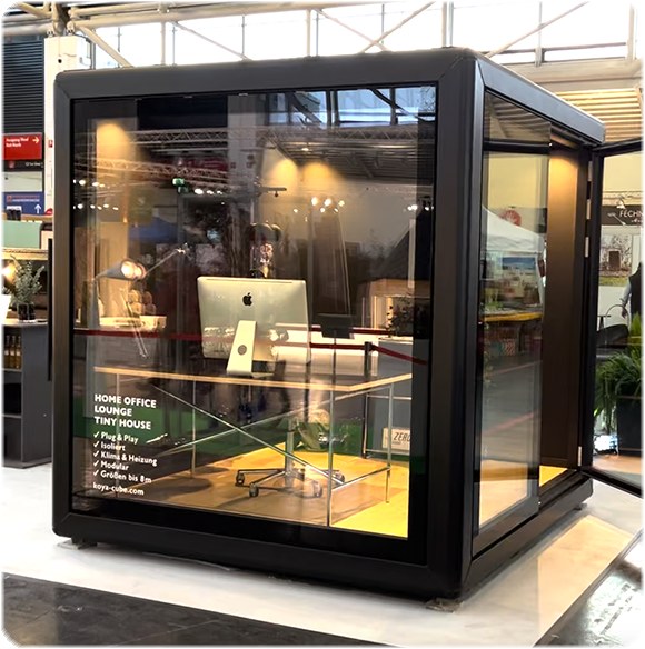 Schwarzer Glaskubus auf einer Messe, innen Schreibtisch mit Monitor, auf der Glasfront „Home Office, Lounge, Tiny House“.