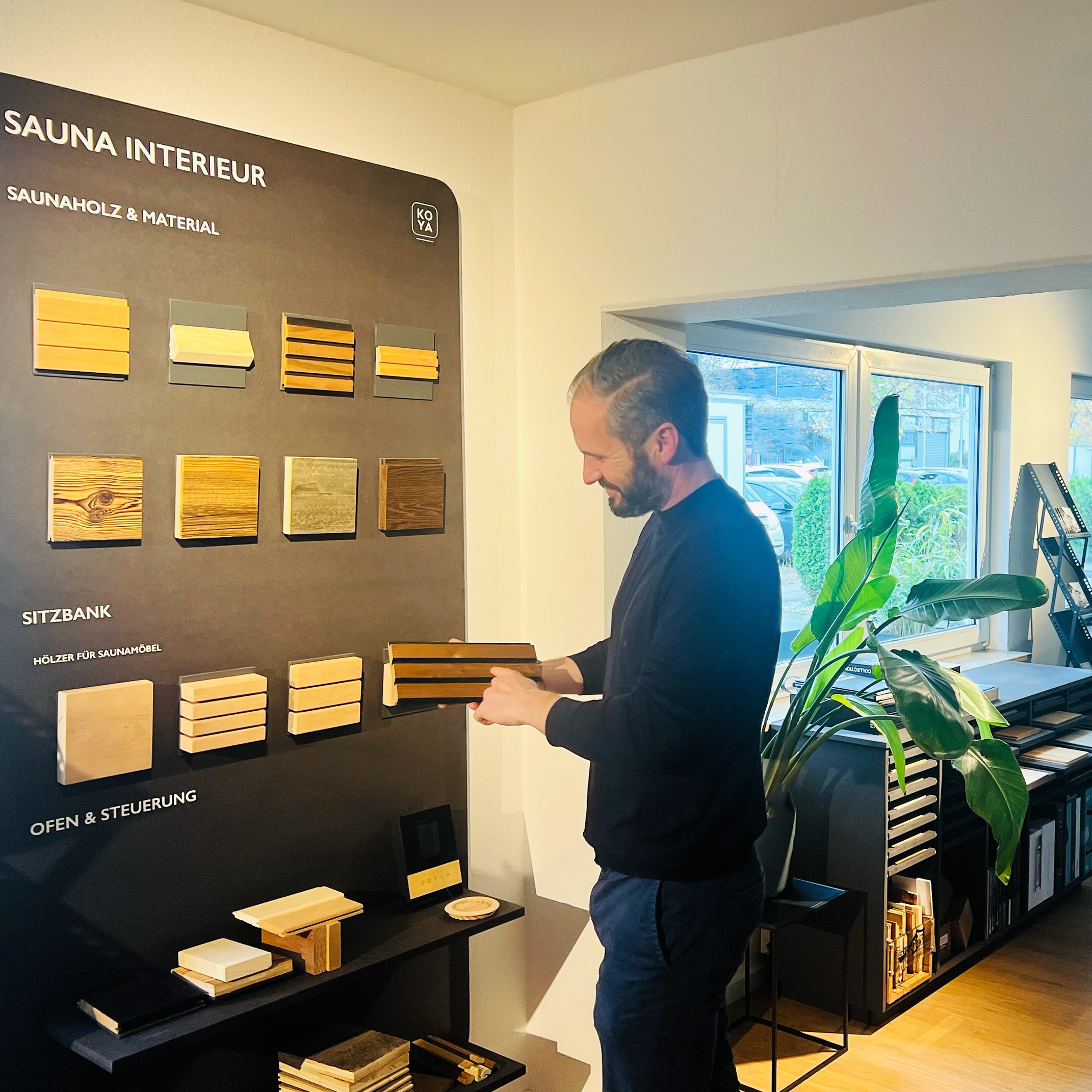 Mann mit Bart hält ein Holzmuster vor einer Musterwand mit Saunaholz, Sitzbank-Hölzern und Ofen-Steuerung.