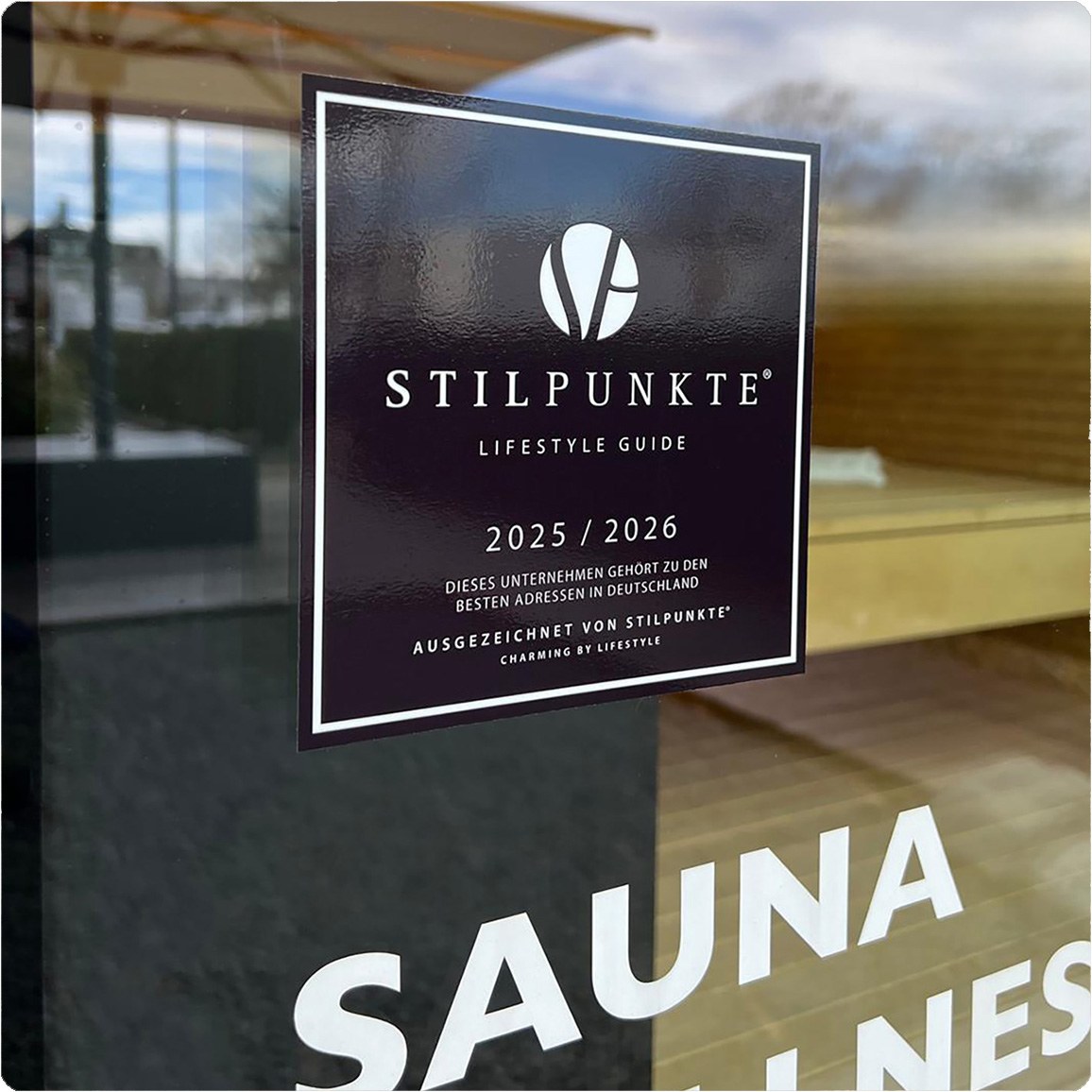 Dunkler Aufkleber „Stilpunkte Lifestyle Guide 2025/2026“ auf einer Glasscheibe, darunter Schriftzug „SAUNA“.