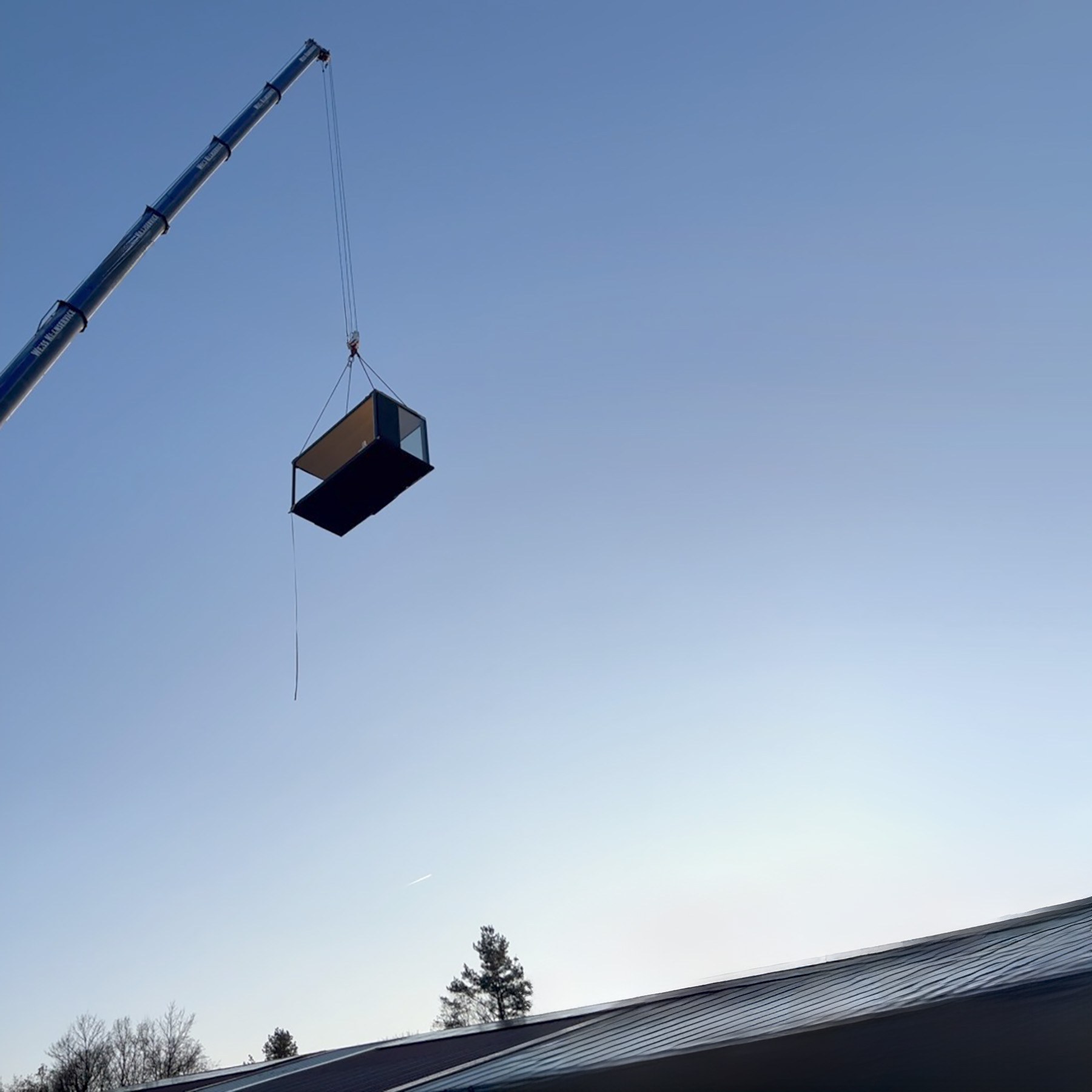 Kran hebt einen dunklen, verglasten Modulbau an Seilen über einem Blechdach vor blauem Himmel.